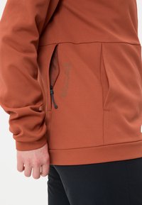 Maglione arancione bruciato in tessuto testurizzato, con tasca laterale con zip e cordino regolabile. Presenta un orlo aderente e polsini comodi.