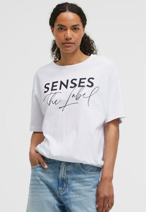 Frau mit lockigem Haar, die ein weißes T-Shirt mit dem Text "SENSES The Label" und hellblaue Jeans trägt, steht mit einer Hand in der Tasche.