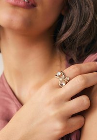 Ring i guld-tone har flettede bånd med klare og frostede sten, sat mod en lys pink stofbaggrund.