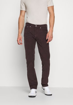 Mand iført mørkebrune manchesterbukser, beige kortærmet skjorte og hvide sneakers, stående mod en ensfarvet hvid baggrund.