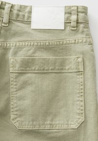 Un pantalon en denim vert clair présente une poche arrière surpiquée et une étiquette blanche carrée avec un design en relief au niveau de la taille.