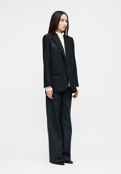 Γυναίκα με μαύρο υπερμεγέθες σακάκι με κεντημένες λεπτομέρειες, λευκό ζιβάγκο, φαρδιά σκούρα παντελόνια και μαύρα μυτερά παπούτσια, όρθια στραμμένη προς τα αριστερά.