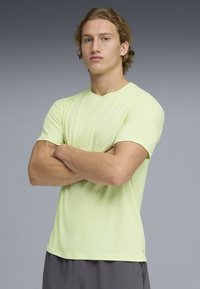 Νεαρός άνδρας με σγουρά μαλλιά, φορώντας αθλητική μπλούζα Puma ανοιχτού κίτρινου χρώματος και γκρι σορτς, στέκεται με σταυρωμένα χέρια μπροστά σε γκρι φόντο.