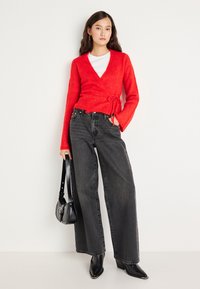 Červený svetr z měkkého pleteného materiálu, kombinovaný s černými širokými kalhotami s vysokým pasem. Doplňky zahrnují malou černou kabelku a boty na podpatku.