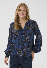 Donna sorridente con lunghi capelli biondi, che indossa una camicetta blu a fiori con bottoni e jeans blu scuro, su uno sfondo neutro.