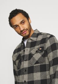 Black and gray checkered flannel shirt with two chest pockets, button closure, rounded collar, and a logo patch on the left pocket.