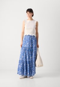 Weißes Smocked-Oberteil mit Rüschensleeves, kombiniert mit einem langen blauen, blumigen Schichtrock. Hält eine hellbeige Tasche und trägt weiße Sneaker.