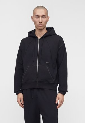 Młody mężczyzna z ogoloną głową, ubrany w czarną bluzę z kapturem na zamek błyskawiczny z przednimi kieszeniami oraz czarne spodnie, na jednolitym szarym tle.