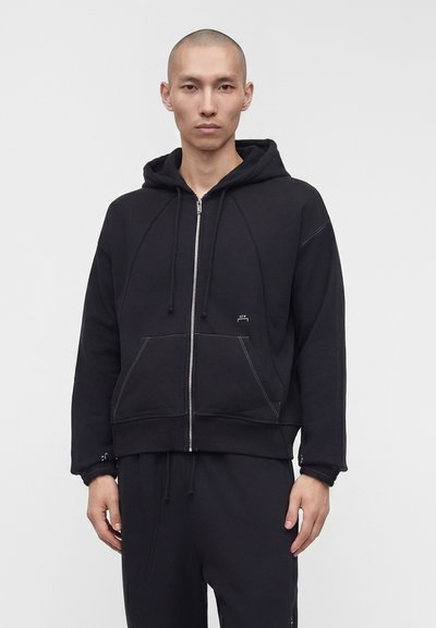 Jeune homme à la tête rasée portant un sweat à capuche zippé noir avec des poches avant et un pantalon noir assorti sur un fond gris uni.