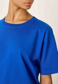 Mėlyna medvilninė trumpa rankovė marškinėliai su apvaliu kaklu, lygiu paviršiumi ir laisvu kirpimu, turintys subtilų surištą apykaklę ir pečių siūles.