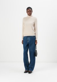Beige genser med høy hals, løse blå jeans, svart veske og svarte spisse sko, stående foran en enkel hvit bakgrunn.