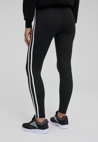 Leggings neri con strisce laterali bianche, realizzati in tessuto elasticizzato, taglio aderente, abbinati a scarpe da ginnastica nere con dettagli in rete.