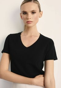 Kobieta o jasnej skórze i spiętych włosach, ubrana w czarną koszulkę z dekoltem w serek, stojąca na tle gładkiej, jasnej ściany, patrząca przed siebie.