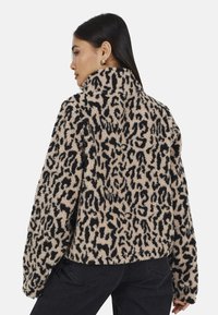 Donna che indossa una giacca pelosa leopardata beige e nera con colletto alto, abbinata a jeans neri, mostrata di spalle e di lato.