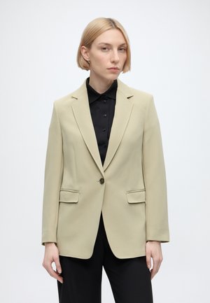 Lyshåret kvinde iført en beige blazer over en sort skjorte med knapper og sorte bukser, står mod en ensfarvet hvid baggrund.