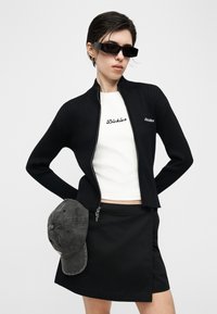 Άτομο που φοράει μαύρο μπουφάν με φερμουάρ, λευκό μπλουζάκι με λογότυπο, μαύρη φούστα, γυαλιά ηλίου, κρατώντας μια γκρι καπέλο πίσω από την πλάτη τους.