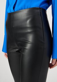 Schwarze Kunstlederhosen mit einem hochschnittigen Design, ausgeführt in einer glatten Textur und mit einem seitlichen Reißverschluss. Kombiniert mit einer blauen Bluse.