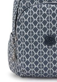 Grå ryggsäck med mönster som har en geometrisk design med vita accenter. Innehåller en cirkulär logotyp-etikett och en silvrig charm som är fäst.