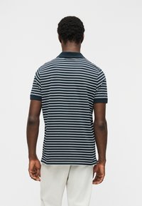 Άνδρας με πλάτη φοράει μπλε ναυτικό και λευκό ριγέ πουκάμισο πόλο με κοντά μανίκια και ανοιχτόχρωμο παντελόνι μπροστά σε απλό φόντο.