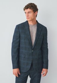 Marintröja med en diskret rutmönster, texturerat tyg, notchkrage och en enkel knappstängning, bärs över en ljusgrå tröja.