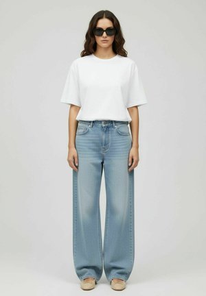 Femme portant des lunettes de soleil noires, un t-shirt blanc surdimensionné, un jean large bleu clair et des ballerines beiges, debout devant un fond uni.