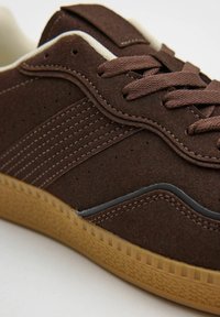 Zapatillas de ante marrón con una superficie texturizada, que cuentan con una suela de goma y detalles en marrón oscuro. Los cordones son del mismo color marrón.