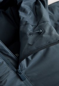 Close-up of black jacket fabric showing zipper, drawstring, and stitching details on smooth, padded material.