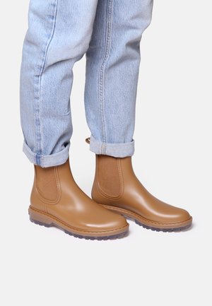 Bottes de cheville en caoutchouc tan avec des panneaux latéraux élastiques, semelles en caoutchouc texturé et accents cousus. Portées avec un jean bleu clair retroussé.