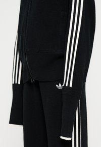 Μαύρο πλεκτό σετ Adidas με λευκές τριπλές ρίγες στα μανίκια και στα παντελόνια, με φερμουάρ και το λογότυπο Adidas trefoil στα παντελόνια.