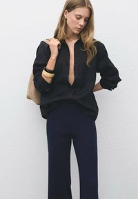 Donna con lunghi capelli biondi che indossa una blusa nera larga, pantaloni a gamba larga blu navy, uno zaino beige e due braccialetti sul polso sinistro, in piedi davanti a un muro bianco.