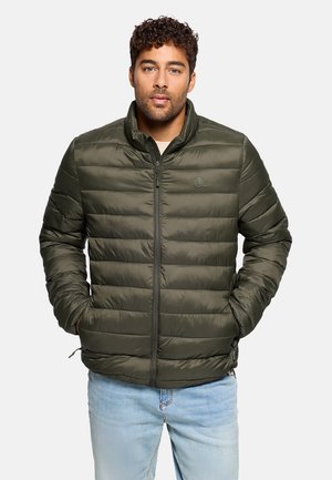 Hombre con cabello rizado, vistiendo una chaqueta acolchada verde oliva con cremallera y vaqueros azul claro, de pie con las manos en los bolsillos sobre un fondo blanco.