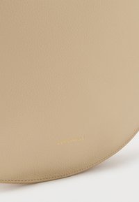 Béžová kožená kabelka s hladkou texturou, zaobleným tvarem a zlatým raženým logem "Coccinelle" v pravém dolním rohu.