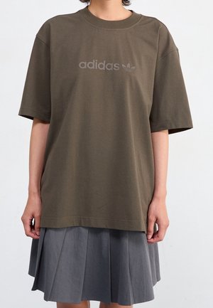 Person iført en oversized olivengrøn Adidas T-shirt med logo og et gråt plisseret knælængde skørt mod en ensfarvet baggrund.