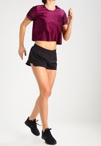 Woman jogging wearing a maroon cropped shirt, black athletic shorts, and black running shoes on a plain background.