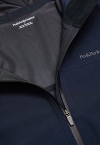 Giacca Peak Performance blu navy con una cerniera nera parzialmente aperta, che mostra il logo del marchio all'interno e all'esterno.
