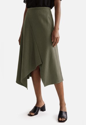 Femme portant une jupe midi asymétrique vert olive avec un détail cache-cœur, associée à des mules noires ouvertes à talon carré.