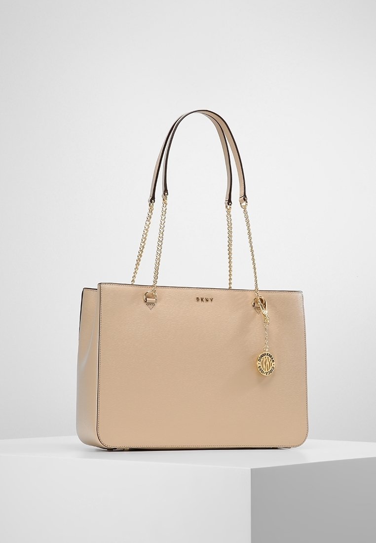 Béžová taška přes rameno vyrobená z texturované kůže, s pozlacenými prvky, dvojitými uchy a přívěskem. Plochý, obdélníkový tvar s otevřeným vrchem.