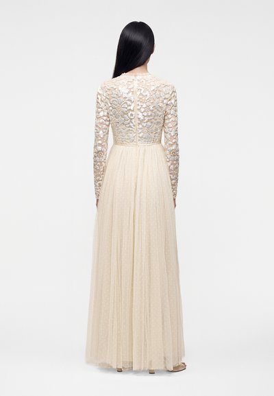 Femme aux longs cheveux noirs portant une robe beige longue jusqu'au sol avec un corsage floral à sequins et une jupe en tulle transparent à pois, vue de dos.