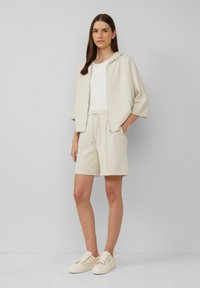 Beige Zip-Hoodie mit kurzen Ärmeln, kombiniert mit passenden beigen Shorts. Darunter ein weißes Shirt und cremefarbene Sneakers mit strukturierten Akzenten.