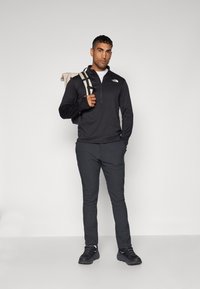 Pull à zip à manches longues noir, pantalon gris, baskets noires et sac à dos beige. Design simple sans motifs ou accents visibles.