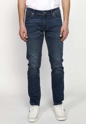 Jeans de mezclilla oscuros ajustados con un ligero desvanecimiento, cinco bolsillos, detalles de costura sutiles, combinados con zapatillas blancas.