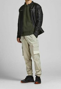 Veste en cuir noir, pull en maille verte, pantalon cargo beige et baskets noires avec accents orange. Matériaux texturés et multiples poches.