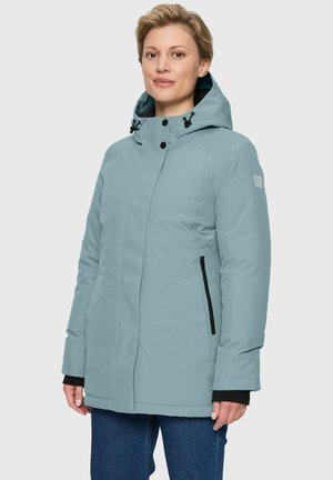 Frau in einer hellblauen Winterjacke mit Kapuze, schwarzen Reißverschlusstaschen und geknöpftem Kragen, steht vor einem schlichten Hintergrund.