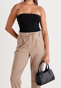 Zwarte strapless top met gegolfde randen, beige taps toelopende broek met trekkoorden, houdt een kleine zwarte handtas met gebogen handvatten vast.