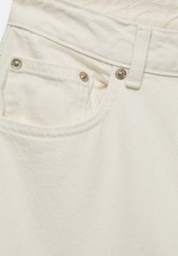 Tissu en denim beige avec une poche avant et des coutures visibles, ainsi que des accessoires en métal argenté. Surface texturée avec un motif diagonal subtil.