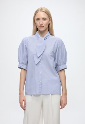 Femme blonde portant un chemisier bleu clair avec des manches courtes bouffantes et un nœud sur le devant, associé à un pantalon blanc à coupe large, debout sur fond blanc.