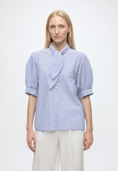 Femme blonde portant un chemisier bleu clair avec des manches courtes bouffantes et un nœud sur le devant, associé à un pantalon blanc à coupe large, debout sur fond blanc.