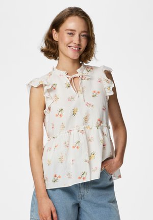 Lächelnde Frau mit kurzen Haaren, die eine weiße ärmellose Bluse mit Blumenmuster und Rüschen sowie hellblaue Jeans trägt, Hand in der Tasche.