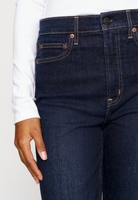 Jeans de mezclilla azul oscuro con herrajes de cobre, costuras amarillas en contraste, y diseño de cinco bolsillos, mostrados a la altura de la cintura media con una blusa blanca de manga larga.