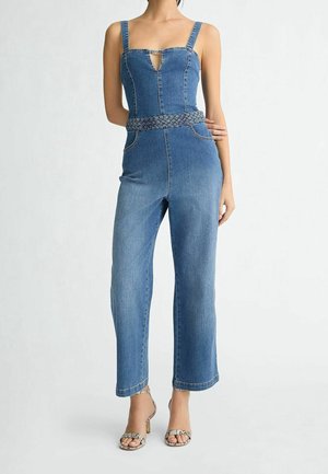 Žena v bezramenném modrém džínovém overalu s tkaným pásem a sandály na vysokém podpatku z hadí kůže, stojící na bílém pozadí.